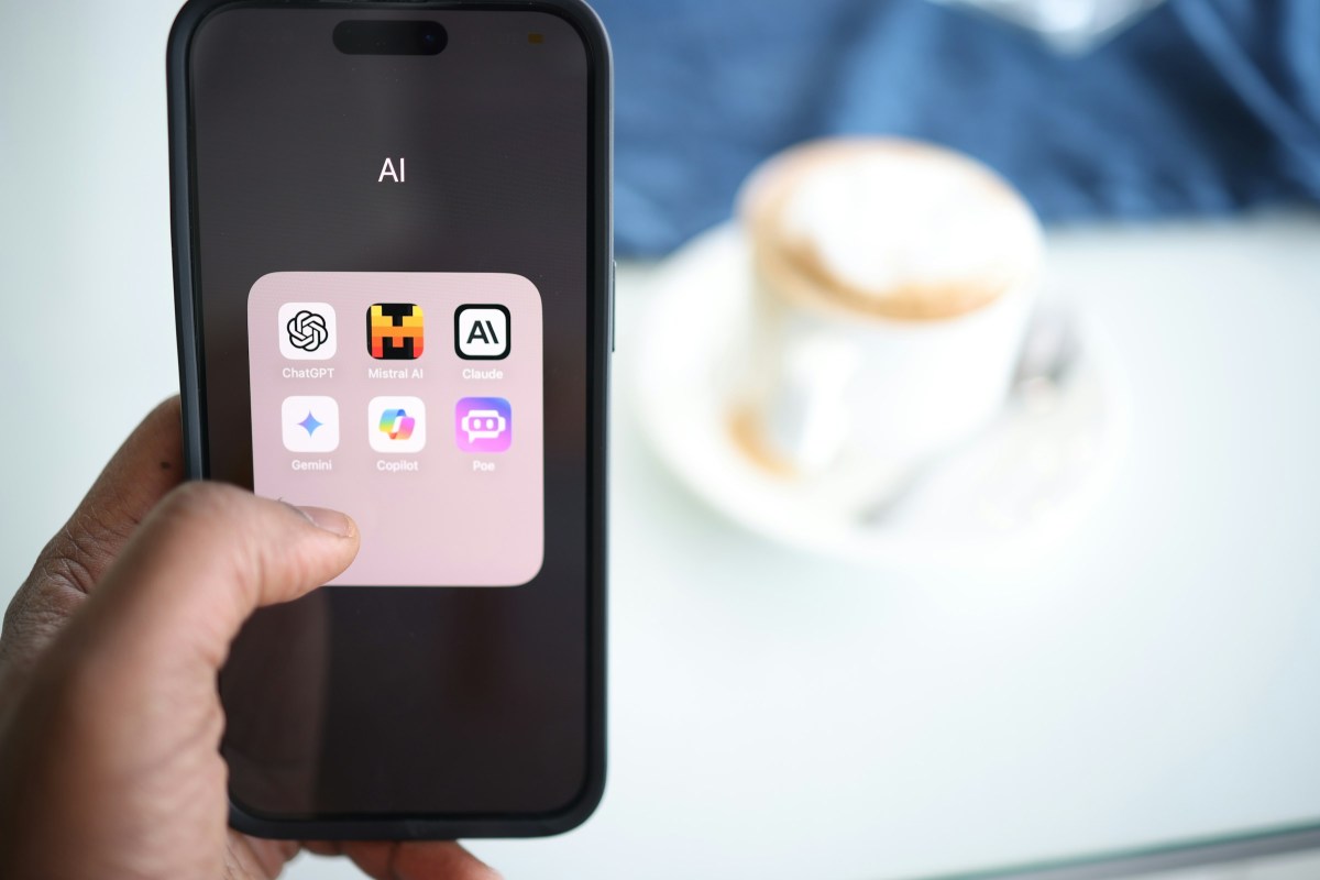1000041296 iPhone held in hand showing a pink rounded app folder labeled AI with six AI app icons (ChatGPT, Mistral AI, Claude, Gemini, Copilot, Poe). Background coffee cup blurred.
