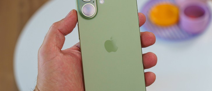 Hand holding an iPhone displaying the home screen with app icons, indoors and slightly blurred background.