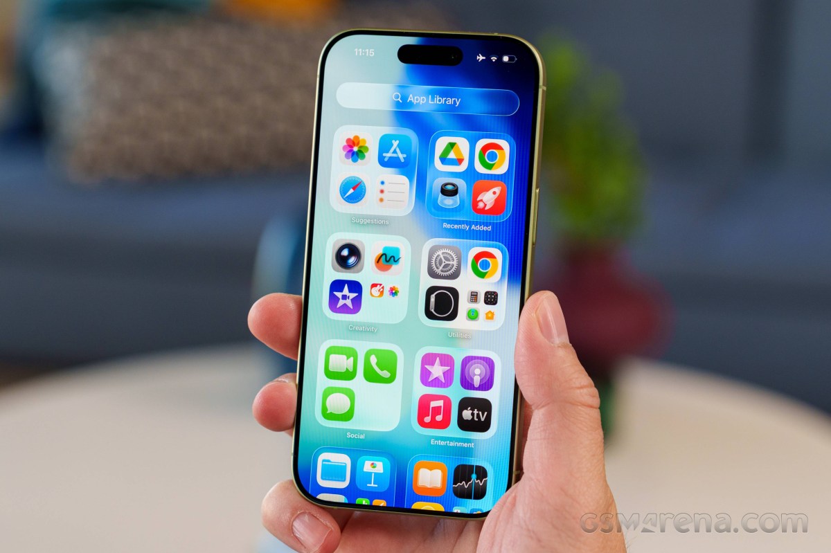 Hand holding an iPhone displaying the home screen with app icons, indoors and slightly blurred background.