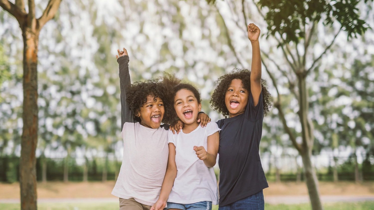 Gentle-strategies-to-build-confidence-in-kids-rooted-rhythm Three smiling children stand arm-in-arm outdoors in a park, raising their fists in celebration.