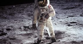 Astronaut in a white space suit on the Moon's surface beside equipment with the lunar lander in the distance.
