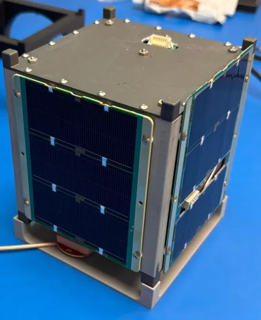 1000047556 Cube satellite prototype with blue solar panels on two sides, mounted on a metal stand on a blue workbench.