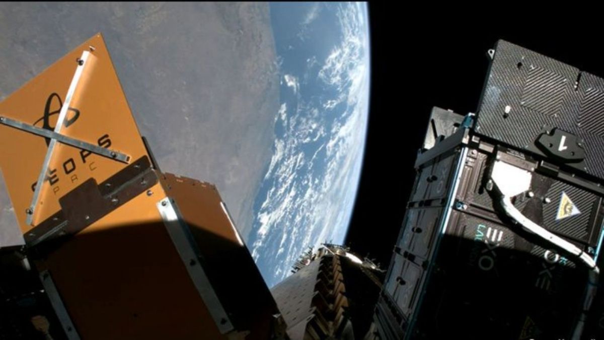 Cube satellite prototype with blue solar panels on two sides, mounted on a metal stand on a blue workbench.