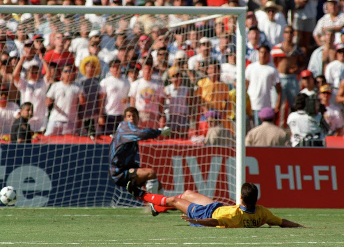 1000049783 Soccer goalkeeper in blue dives to block a shot as a yellow-shirted attacker slides on the grass near the goal. Crowd watches in the background.