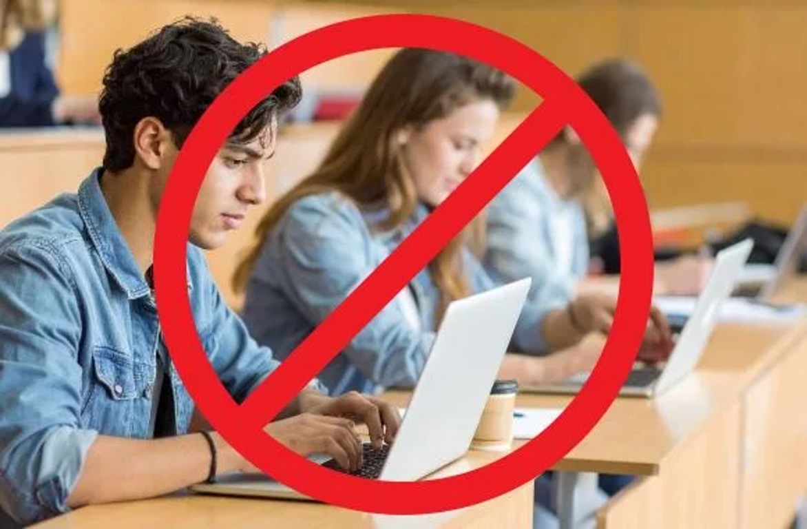 Students in a classroom using laptops with a red no-entry circle, indicating no laptops allowed.