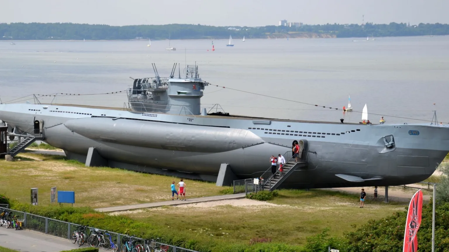 Large gray submarine on display with visitors walking up a stairway to board; water and boats in the background.