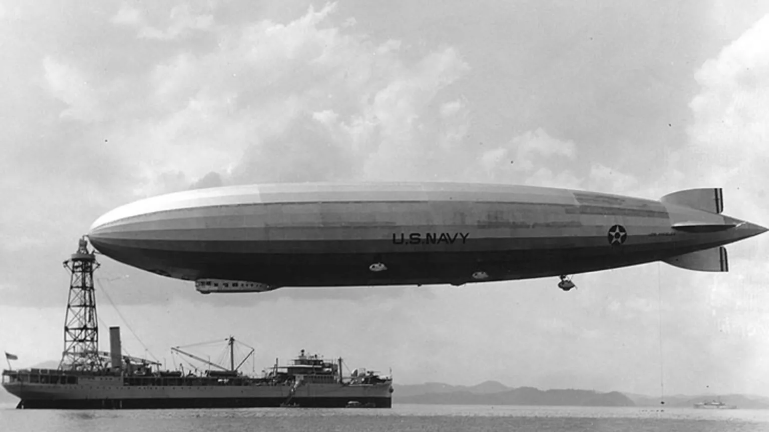 Historic black-and-white photo of a U.S. Navy airship (blimp) hovering near a harbor ship above calm water.