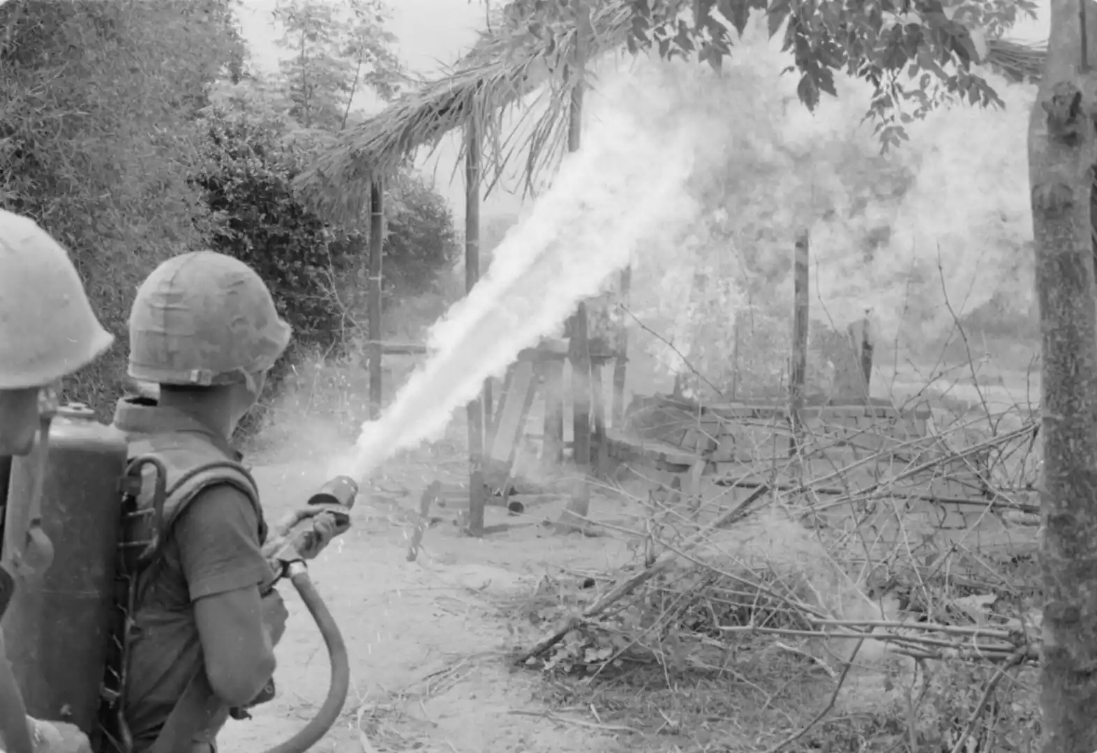 Two soldiers in helmets with backpacks spray water from a hose at a smoky, rubble-filled outdoor scene.