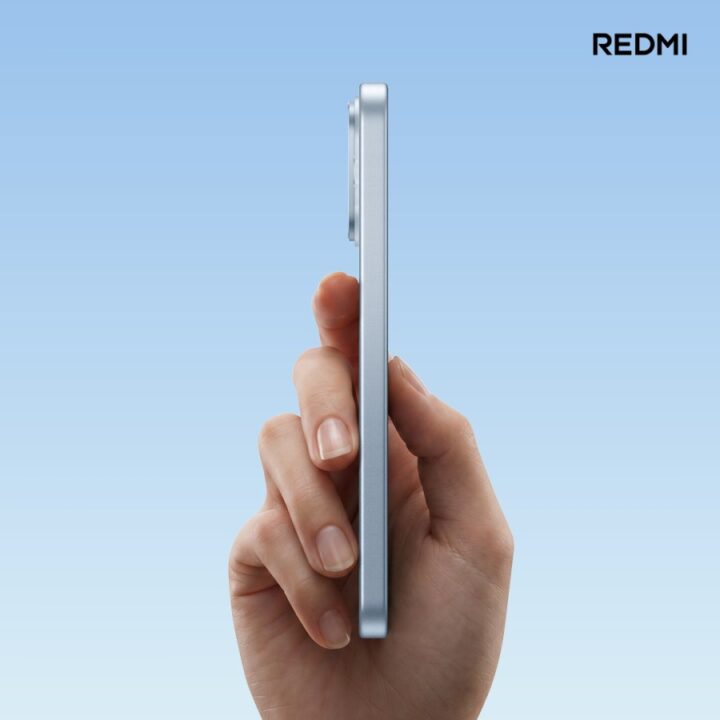 gsmarena_003 Side profile of a hand holding a slim silver Redmi smartphone against a gradient blue background.