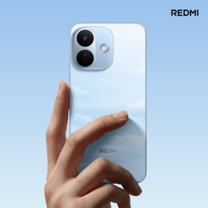 gsmarena_004 Hand holding a light blue Redmi smartphone against a gradient blue background, showcasing the rear camera module and branding on the back.