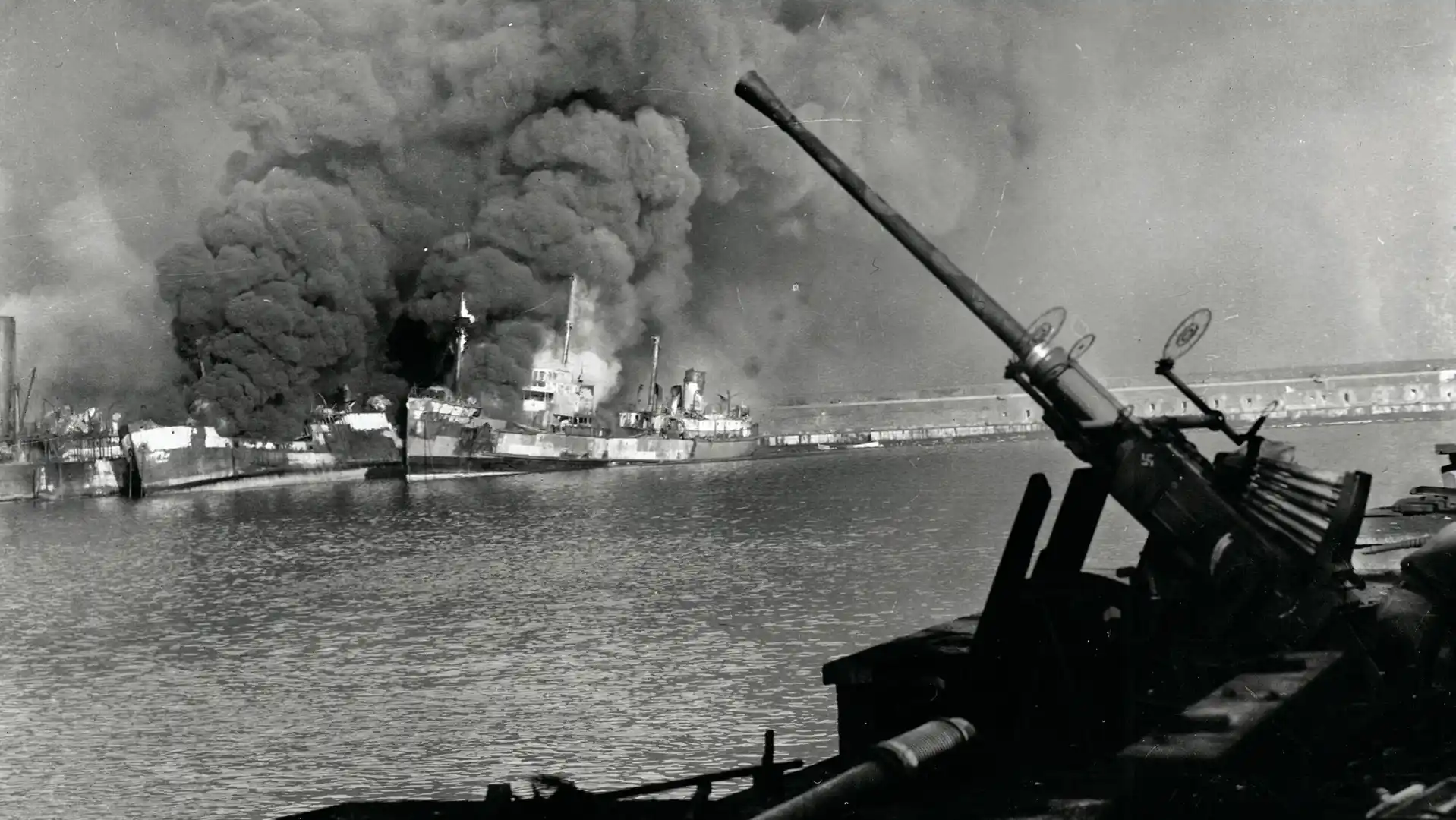 Warship under attack with a massive smoke plume rising from the hull, seen from a foreground naval gun on the water.