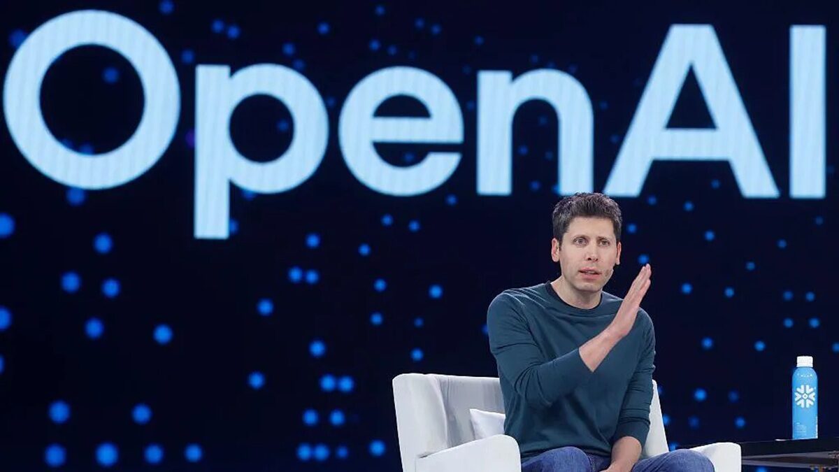 openai Man in a teal shirt sits on a white chair on stage with a large OpenAI sign behind him; a blue bottle sits on a table beside him.